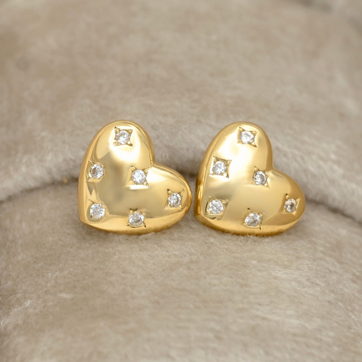 A pair of gold heart-shaped stud earrings, each adorned with small clear  round CZ stones in a star setting, displayed on a soft beige textured surface.