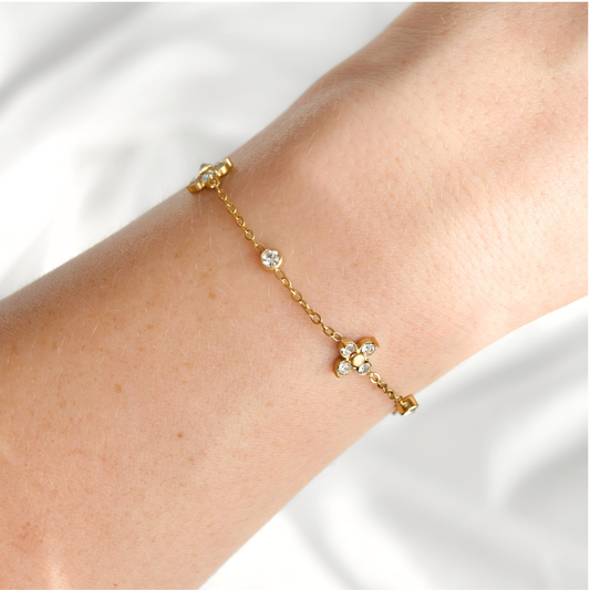 A delicate gold chain bracelet with a small flower motif and clear rhinestones, shown worn on a person's wrist against a soft white fabric background.