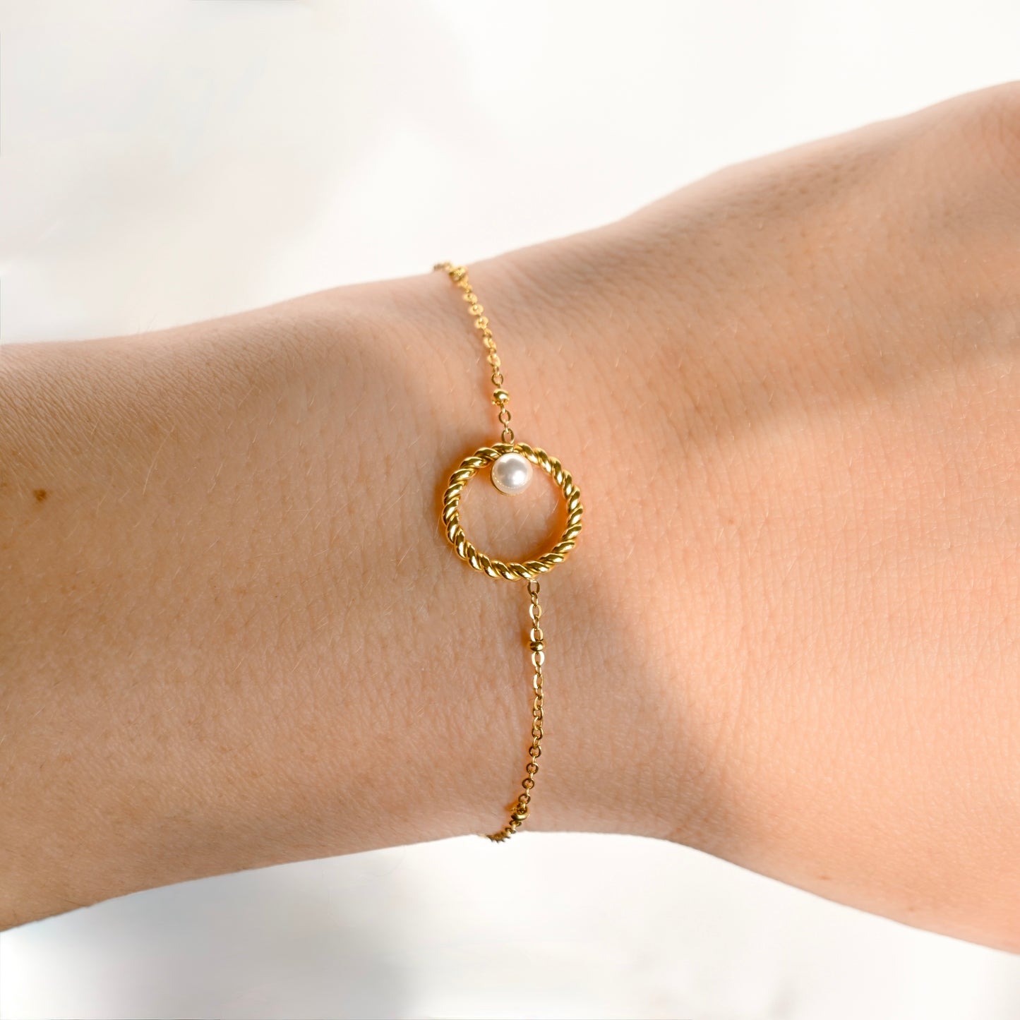 A delicate gold chain bracelet with a twisted circular ring and a single white pearl, shown worn on a person's wrist.