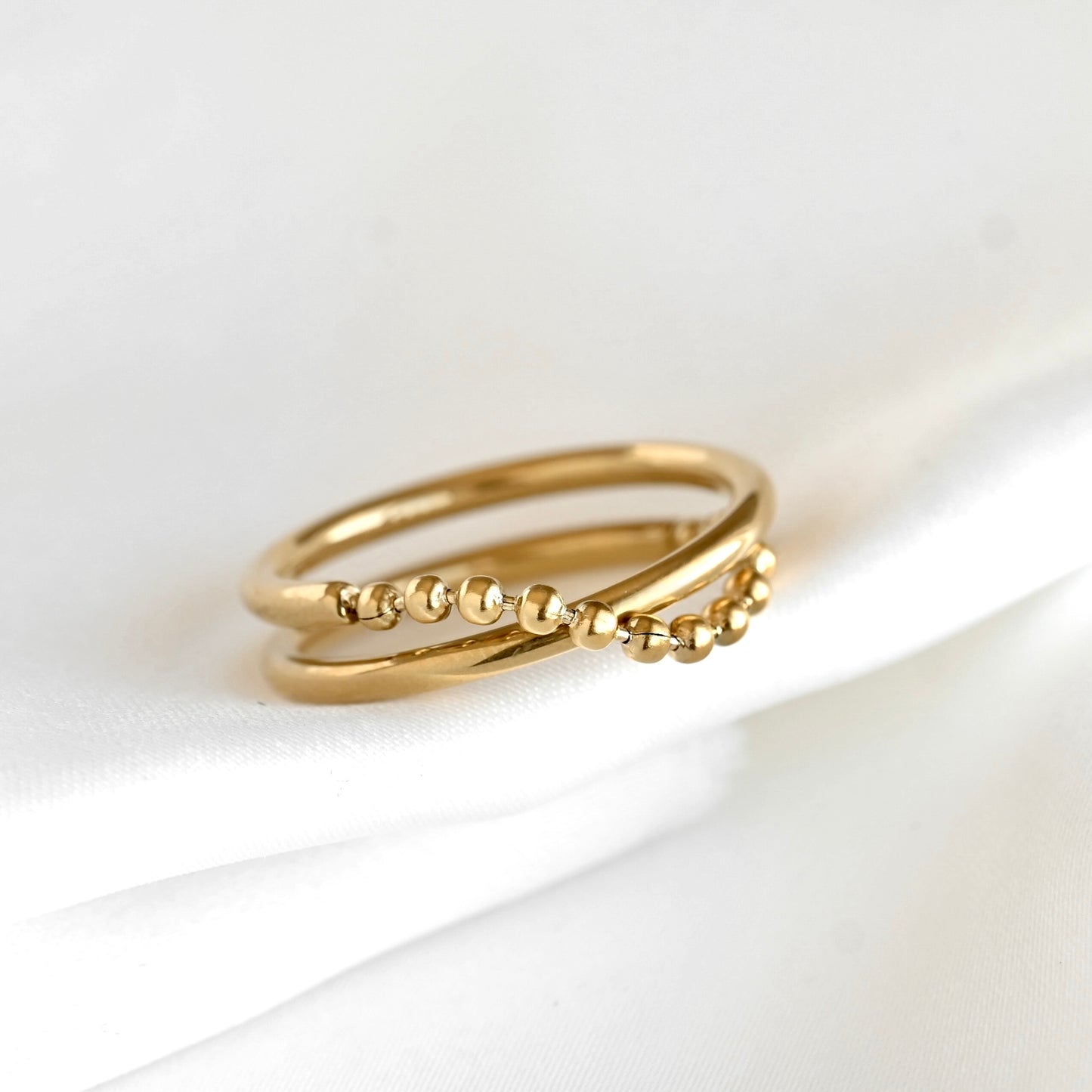A delicate gold double ring with small bead chain details, resting on a soft white fabric background.