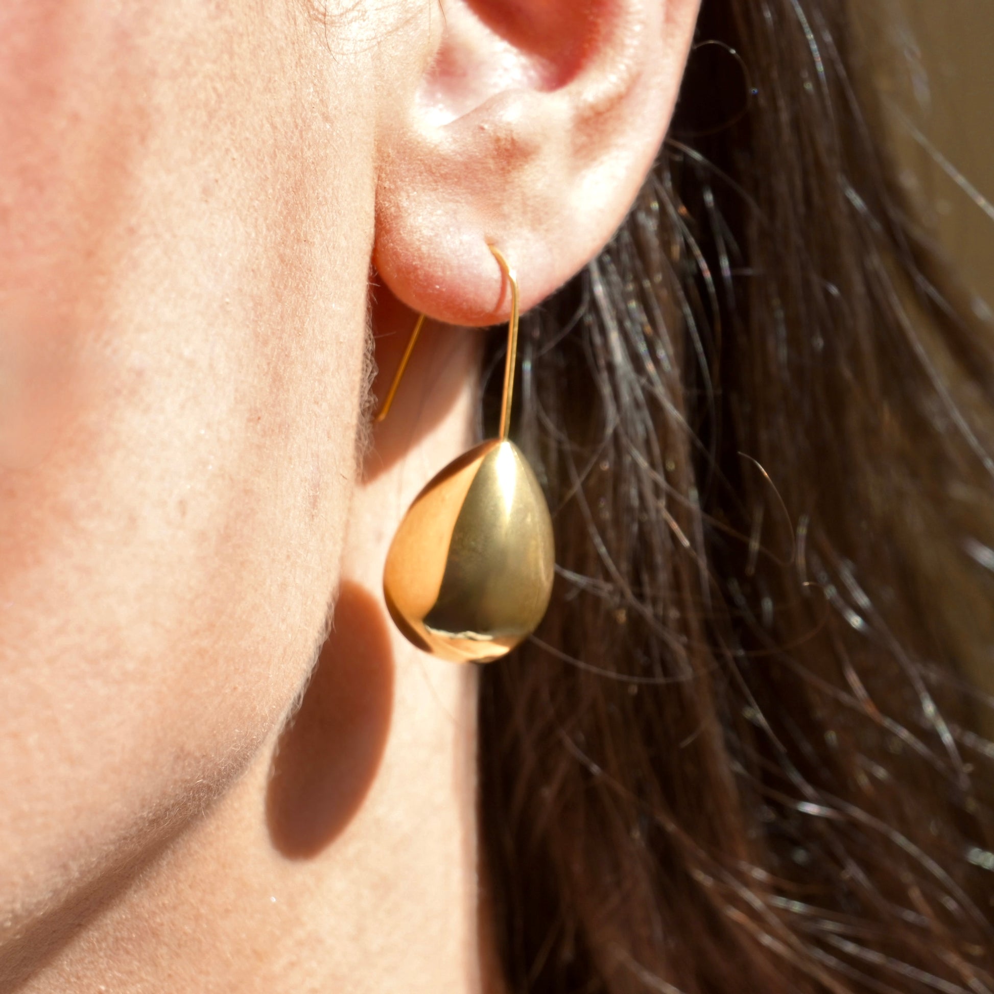 Gold teardrop earring worn on an ear with a blurred background under direct sunlight