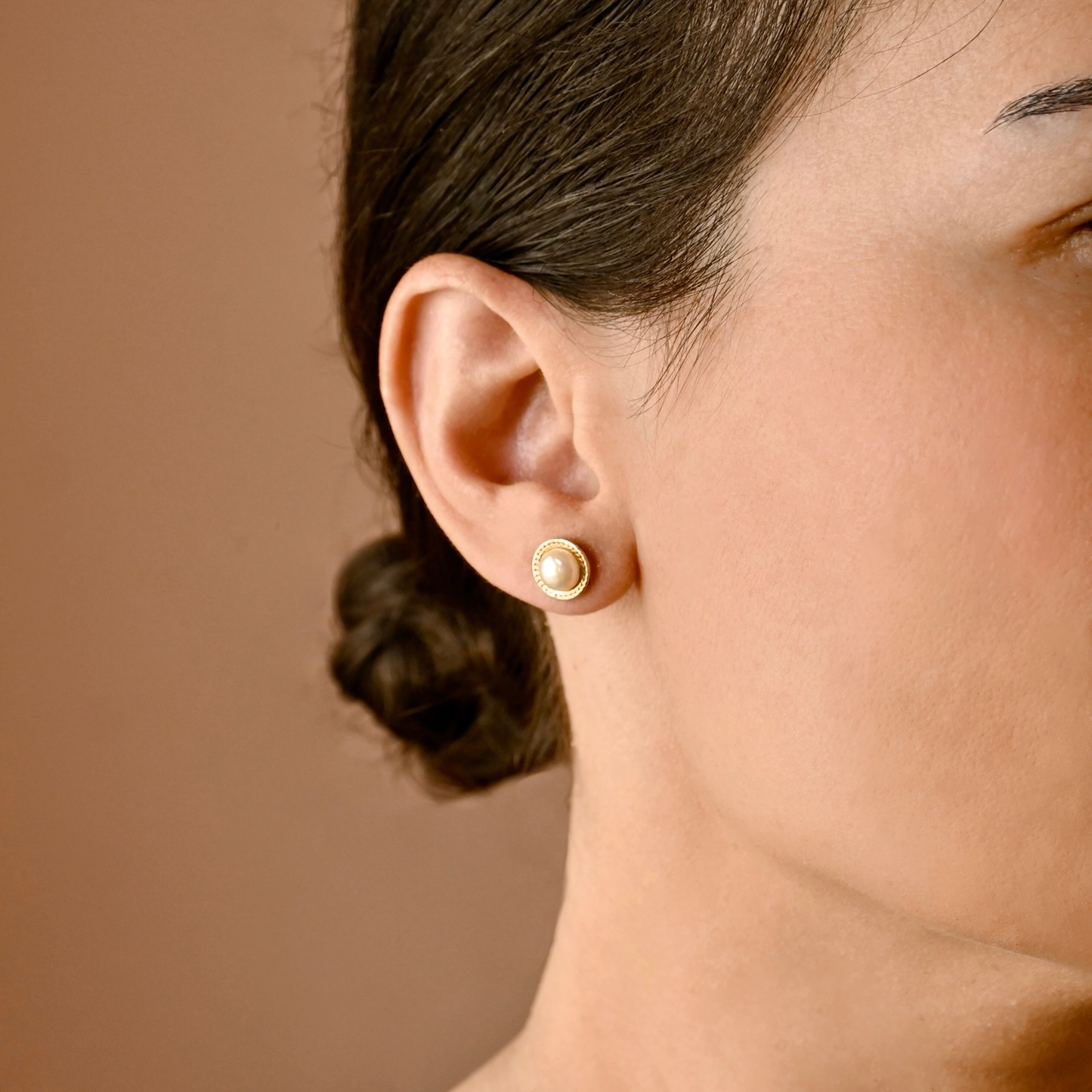 A close-up of a woman's ear wearing a small round gold stud earring with a white pearl center, against a warm brown background.