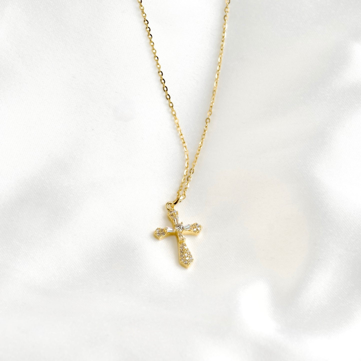 A delicate gold cross pendant necklace adorned with small clear crystals, resting on a soft, white satin fabric background.