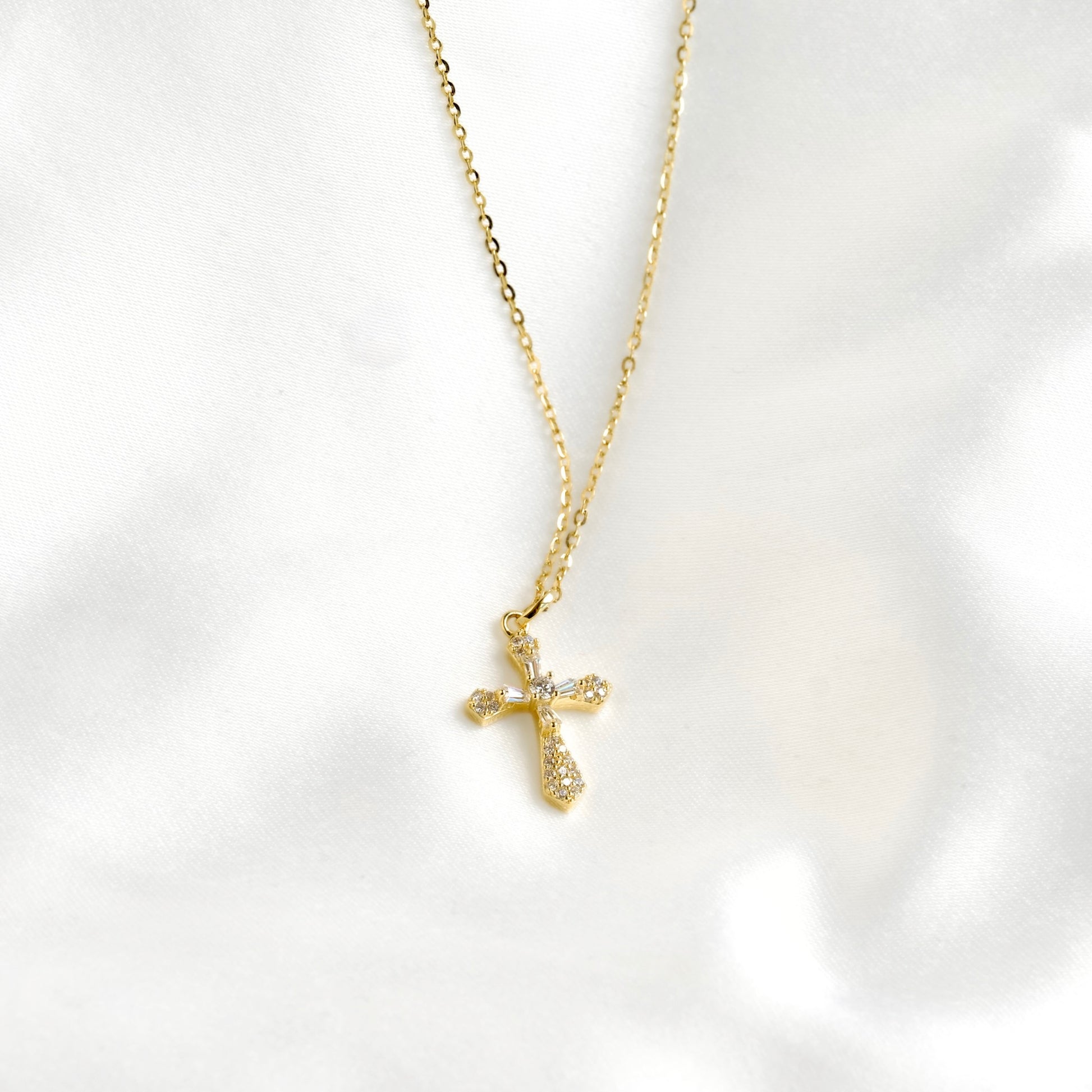 A delicate gold cross pendant necklace adorned with small clear crystals, resting on a soft, white satin fabric background.