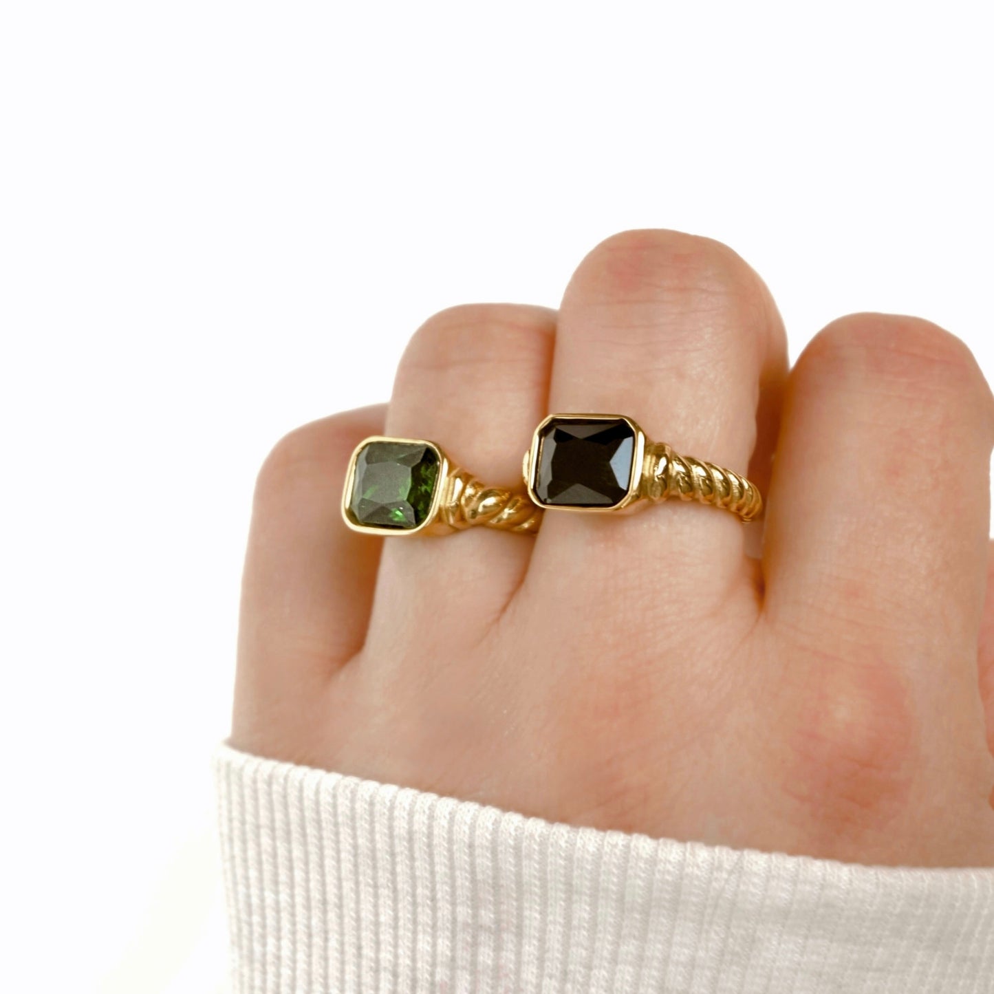 A close-up of a hand wearing two gold rings with square-cut stones, one green and one dark jet black, against a clean white background.