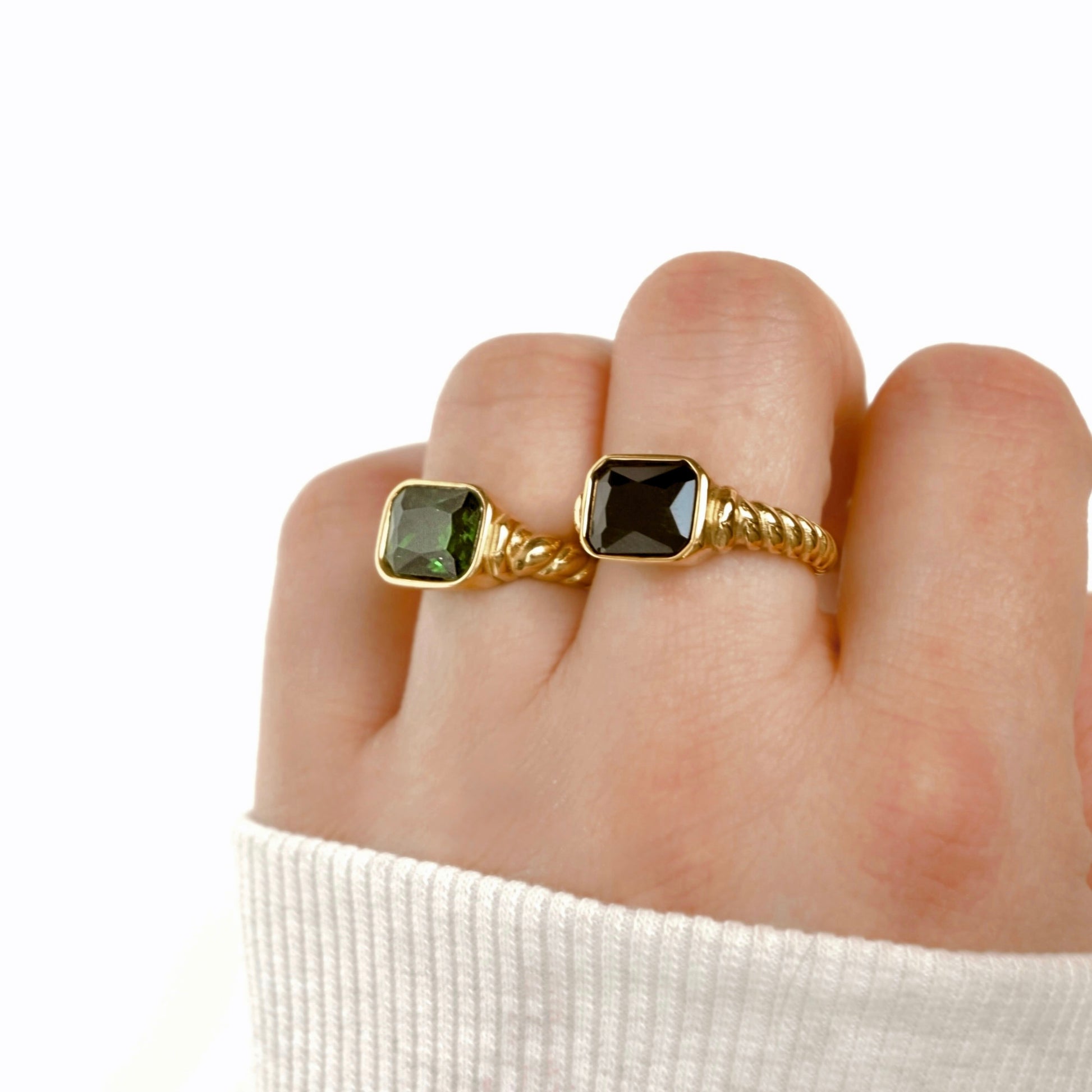 A close-up of a hand wearing two gold rings with square-cut stones, one green and one dark jet black, against a clean white background.