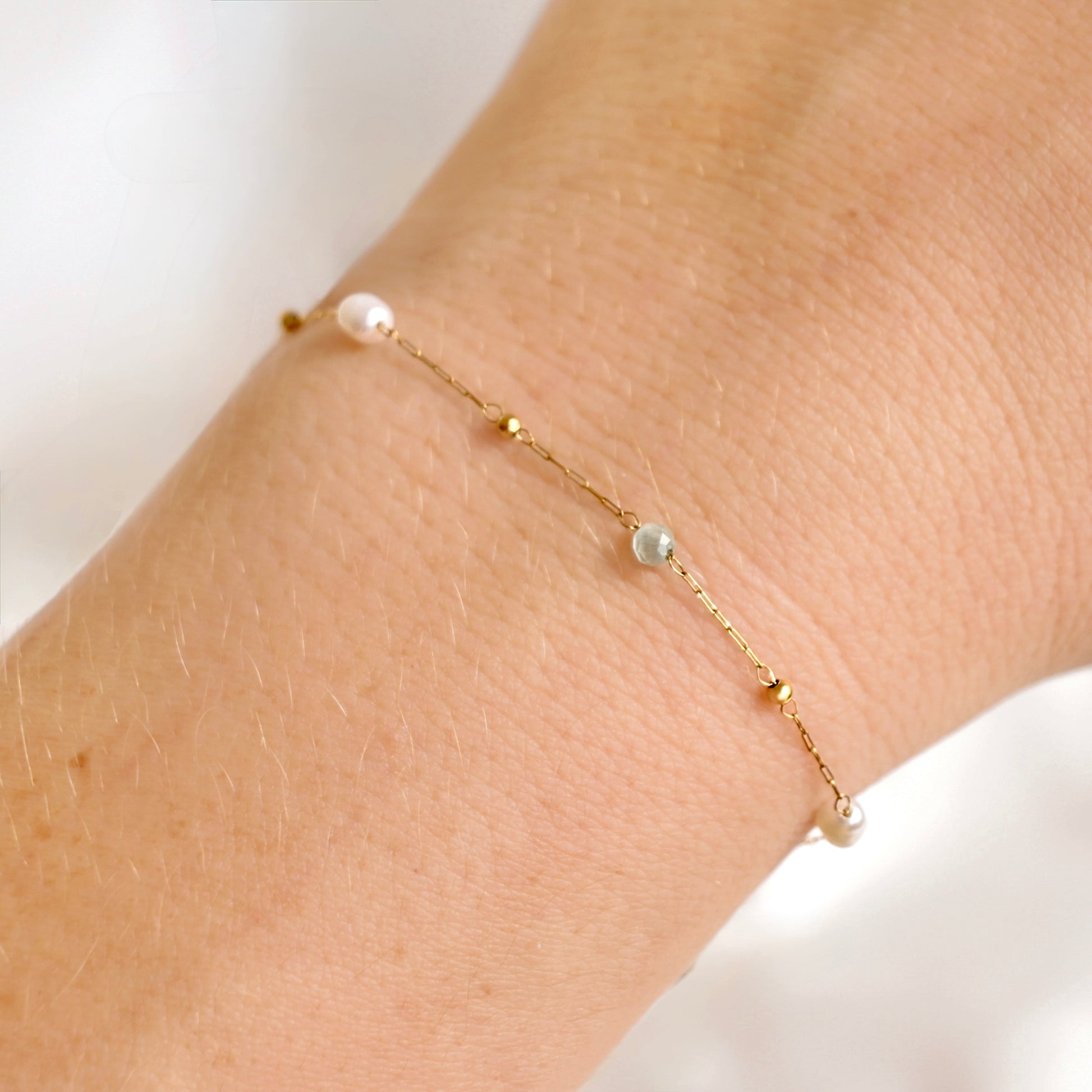 A delicate gold chain bracelet with small white pearls and a pale blue bead, shown worn on a person's wrist.