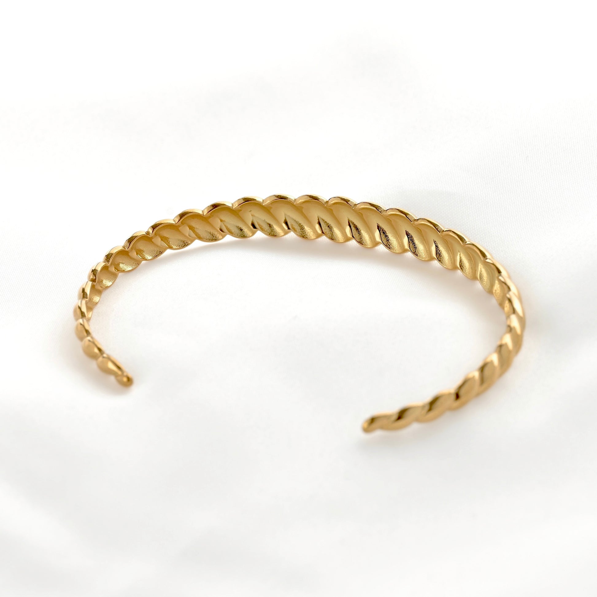 A gold-colored twisted open cuff bracelet with a textured, rope-like spiral design, displayed on a soft white fabric background.