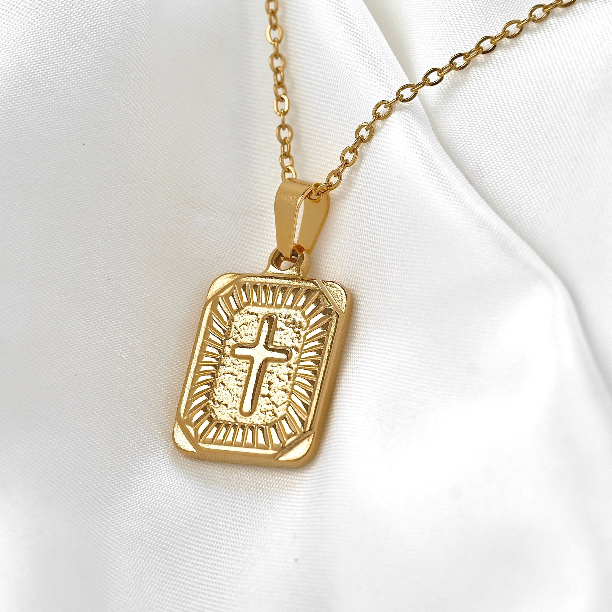 A gold rectangular pendant necklace featuring an engraved cross in the center, displayed on a soft white satin fabric background.