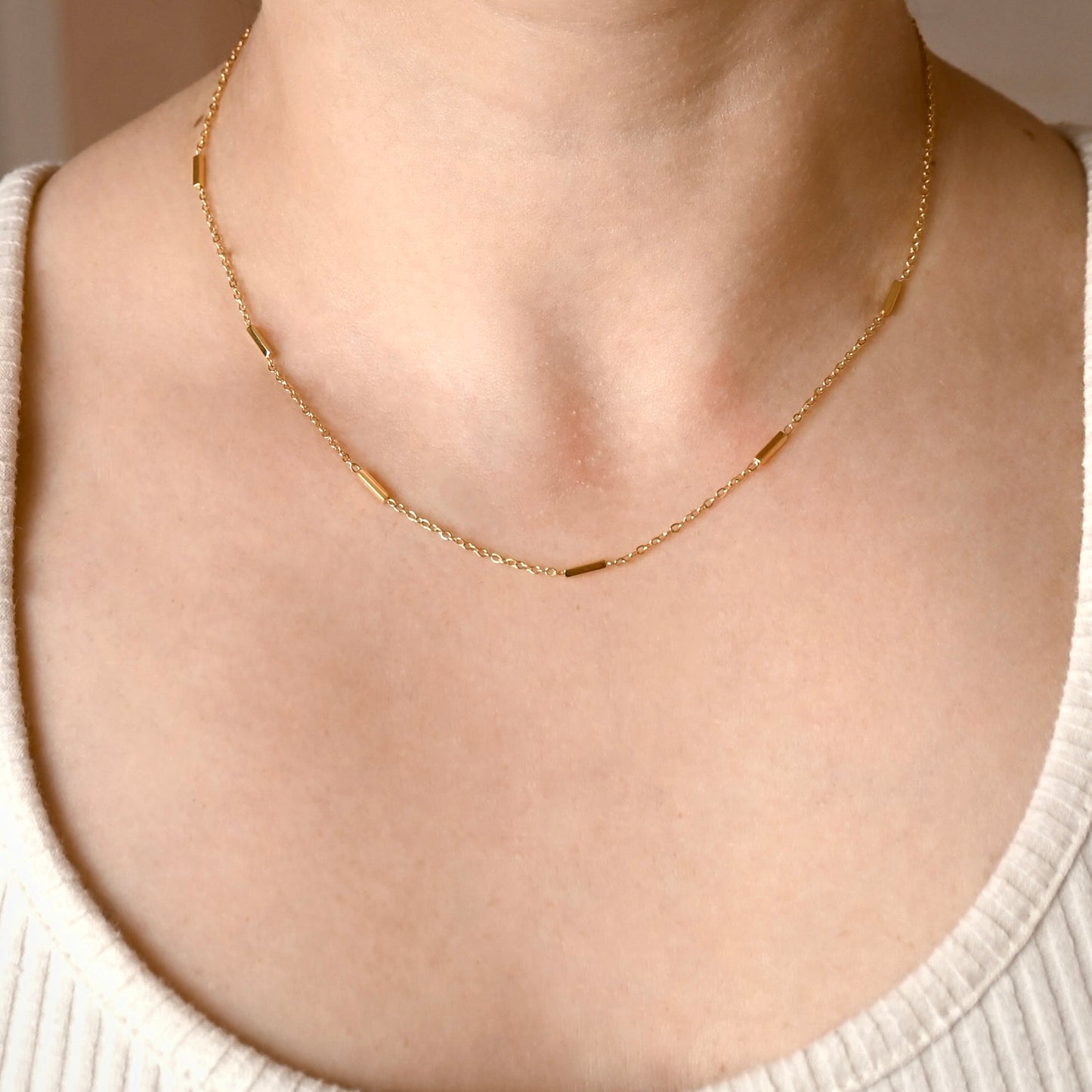 A delicate gold chain necklace with small polished rectangular links, worn on a model's neck over a cream-colored ribbed top.