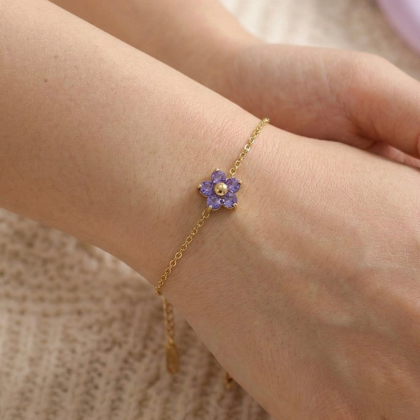 Gold bracelet with a purple flower charm on a wrist against a neutral background