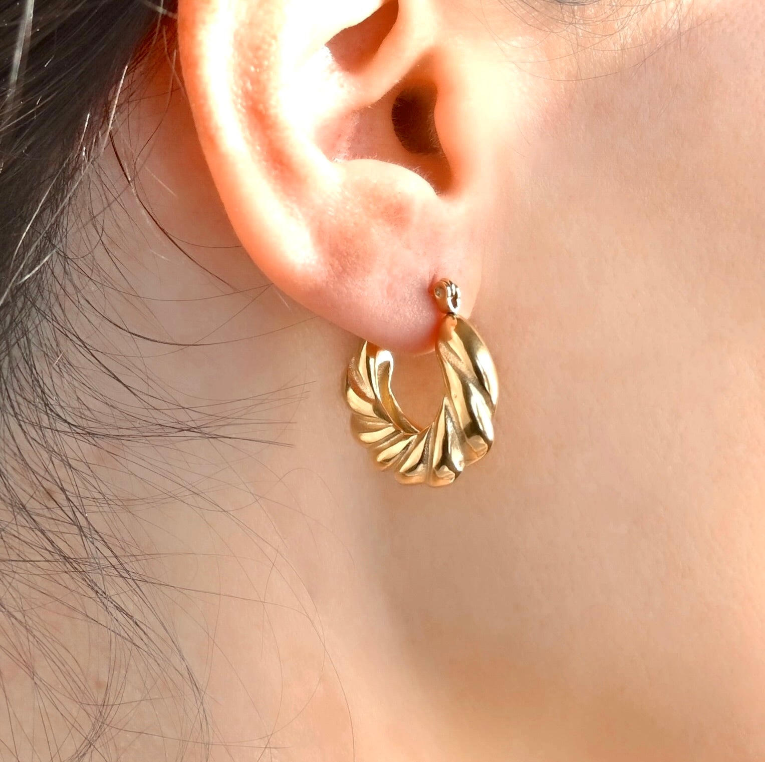 Bold gold twisted croissant hoop earrings worn on an ear with a blurred background