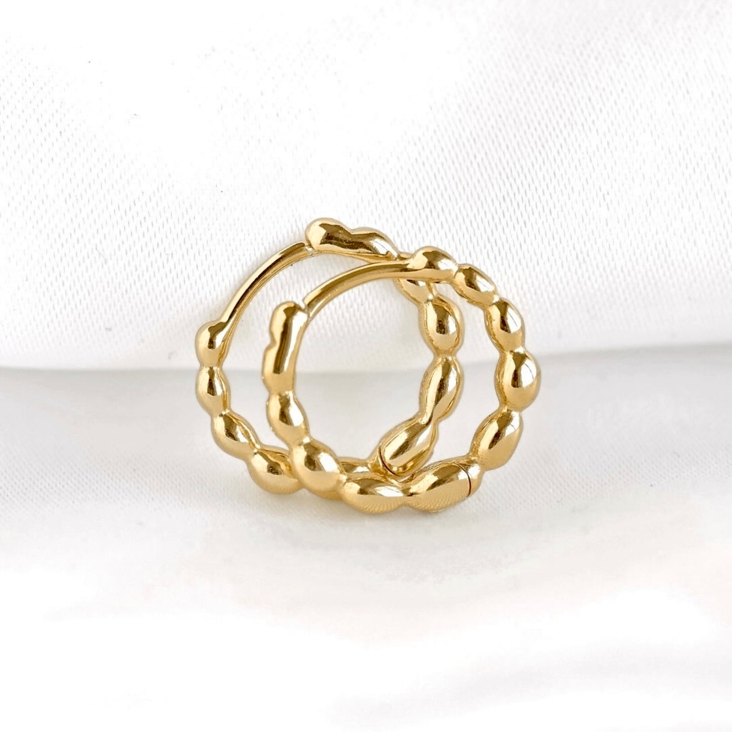 A par of gold beaded huggie hoops with a textured, beaded circular design, displayed on a soft white fabric background.