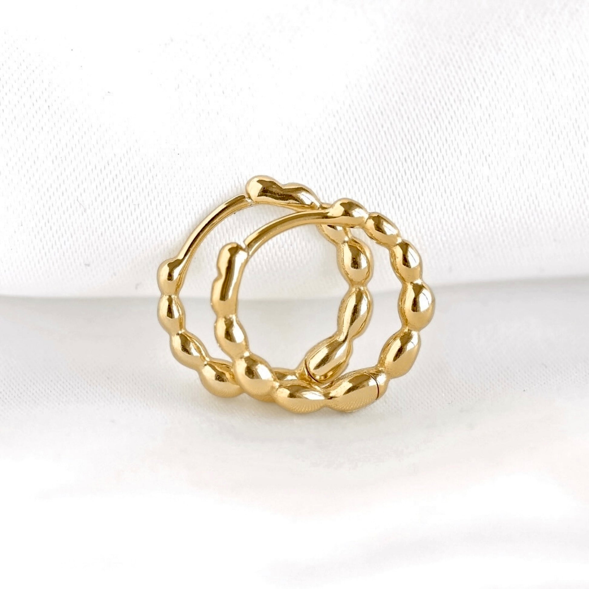 A par of gold beaded huggie hoops with a textured, beaded circular design, displayed on a soft white fabric background.