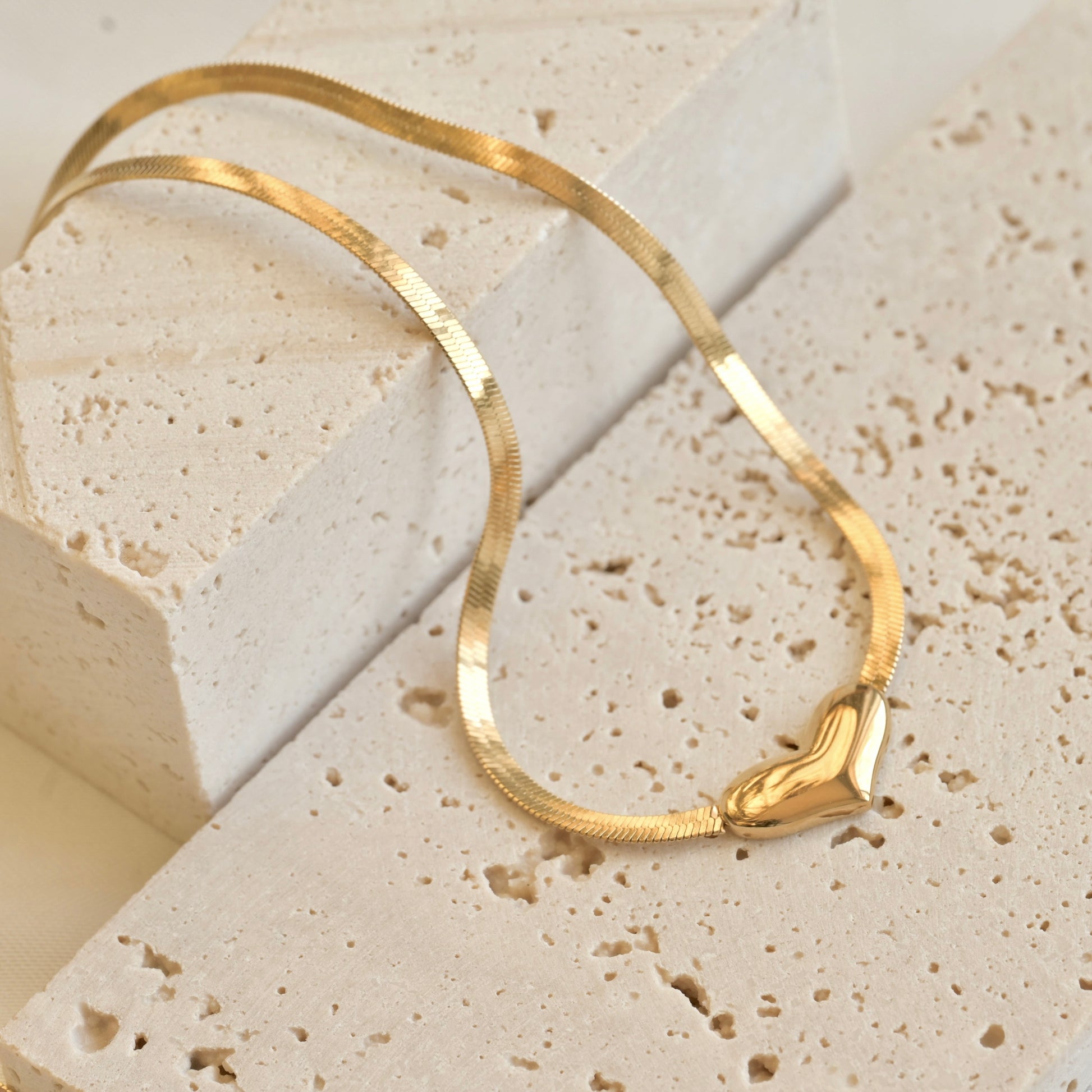 Close-up of a gold choker necklace wit flat heart centerpiece on a textured beige surface