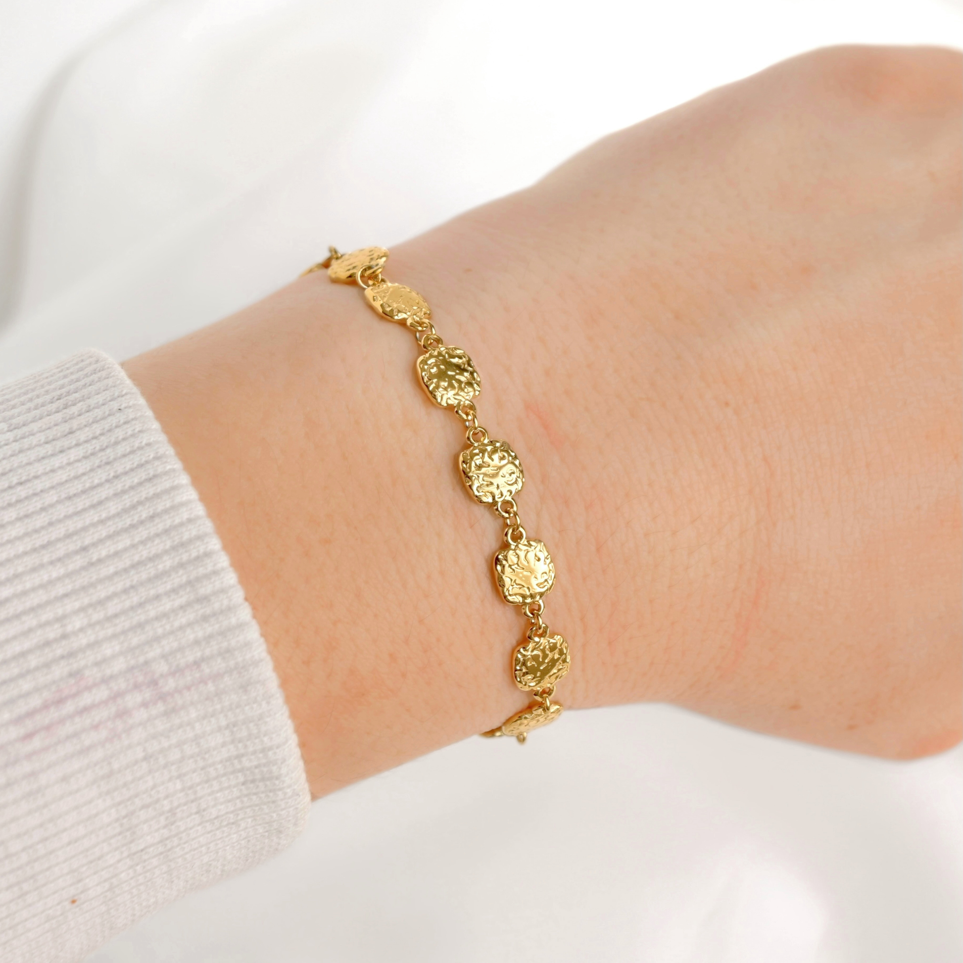 A delicate gold bracelet with textured, rounded oval links worn on a person's wrist, paired with a white ribbed knit sleeve.
