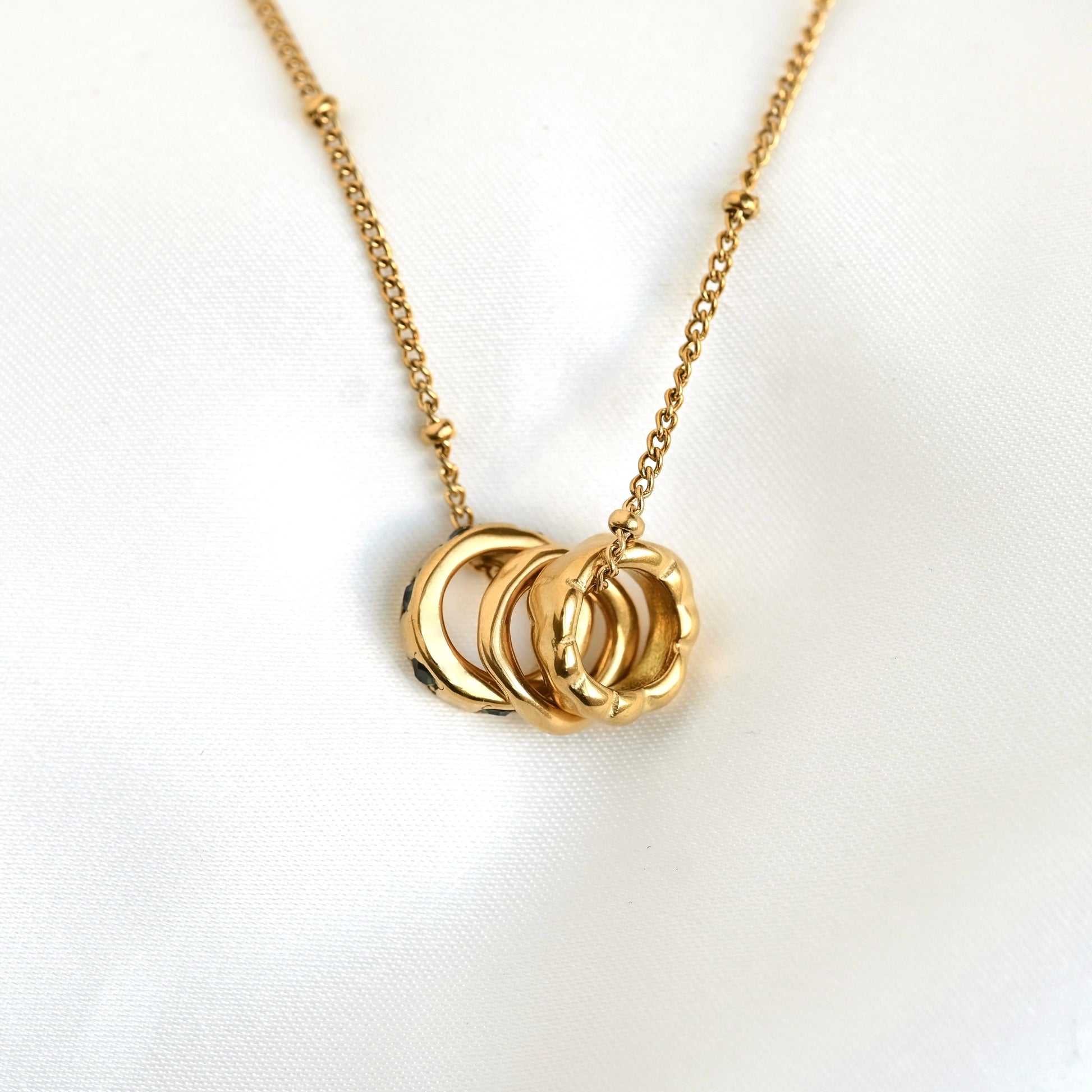 A gold chain necklace with a pendant made of three interlocking textured rings, displayed on a white fabric background.