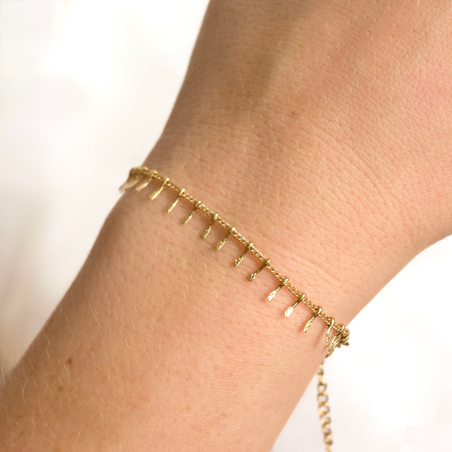 A delicate gold chain bracelet with small dangling spikes, shown worn on a person's wrist against a bright white background.
