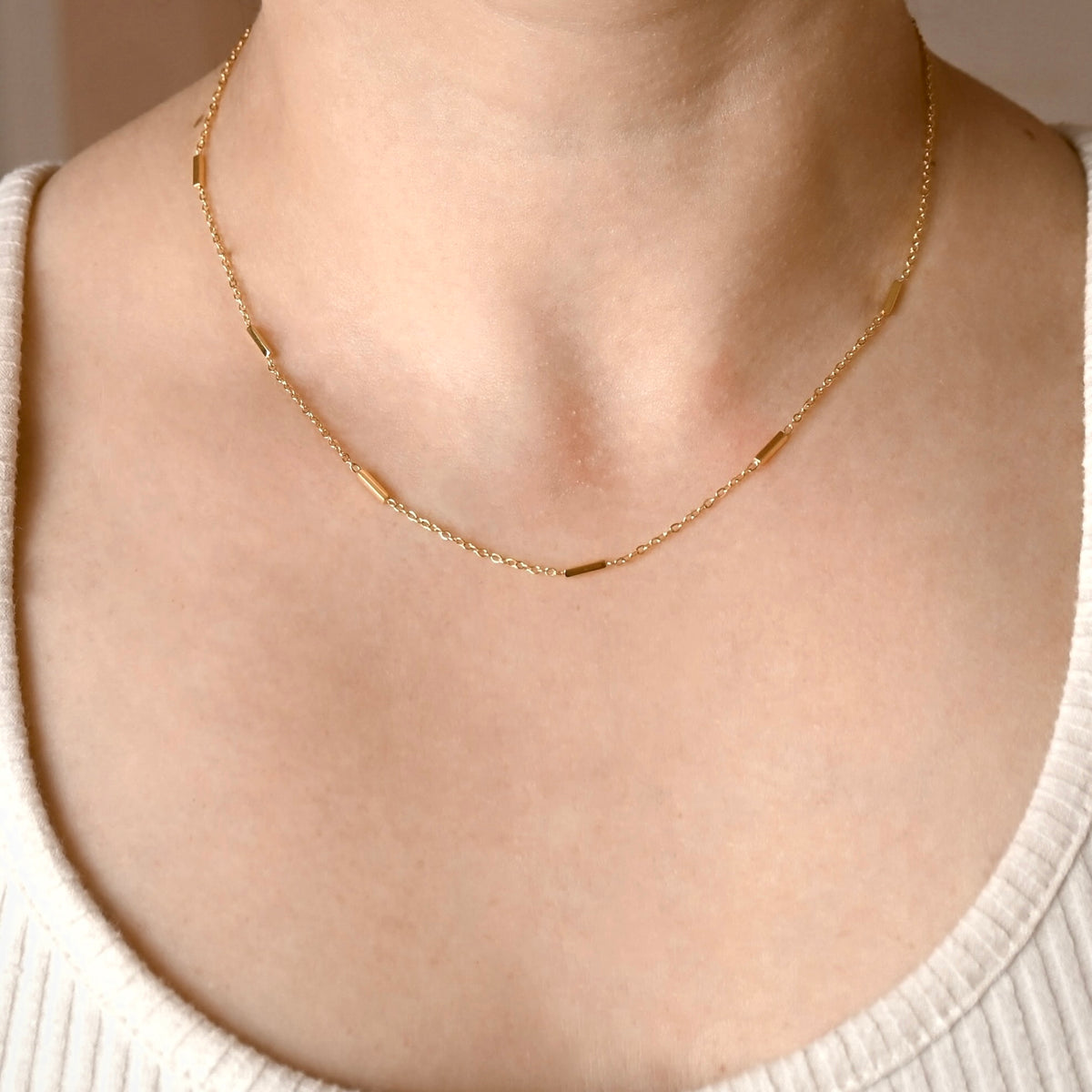 A delicate gold chain necklace with small cylindrical links, worn on a person's neck over a cream-colored ribbed top.