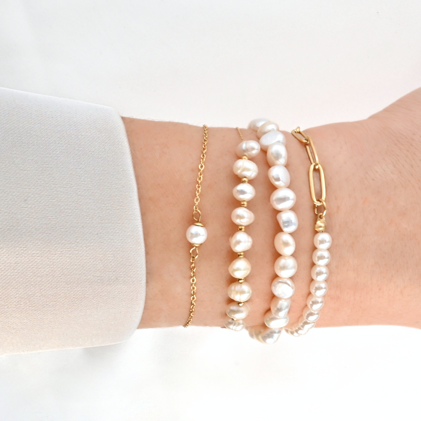 Side view of a wrist wearing four pearl bracelets including single pearl, pearls with gold spacers, full pearl and simulated pearls with paperclip chain designs on a wrist with a white background