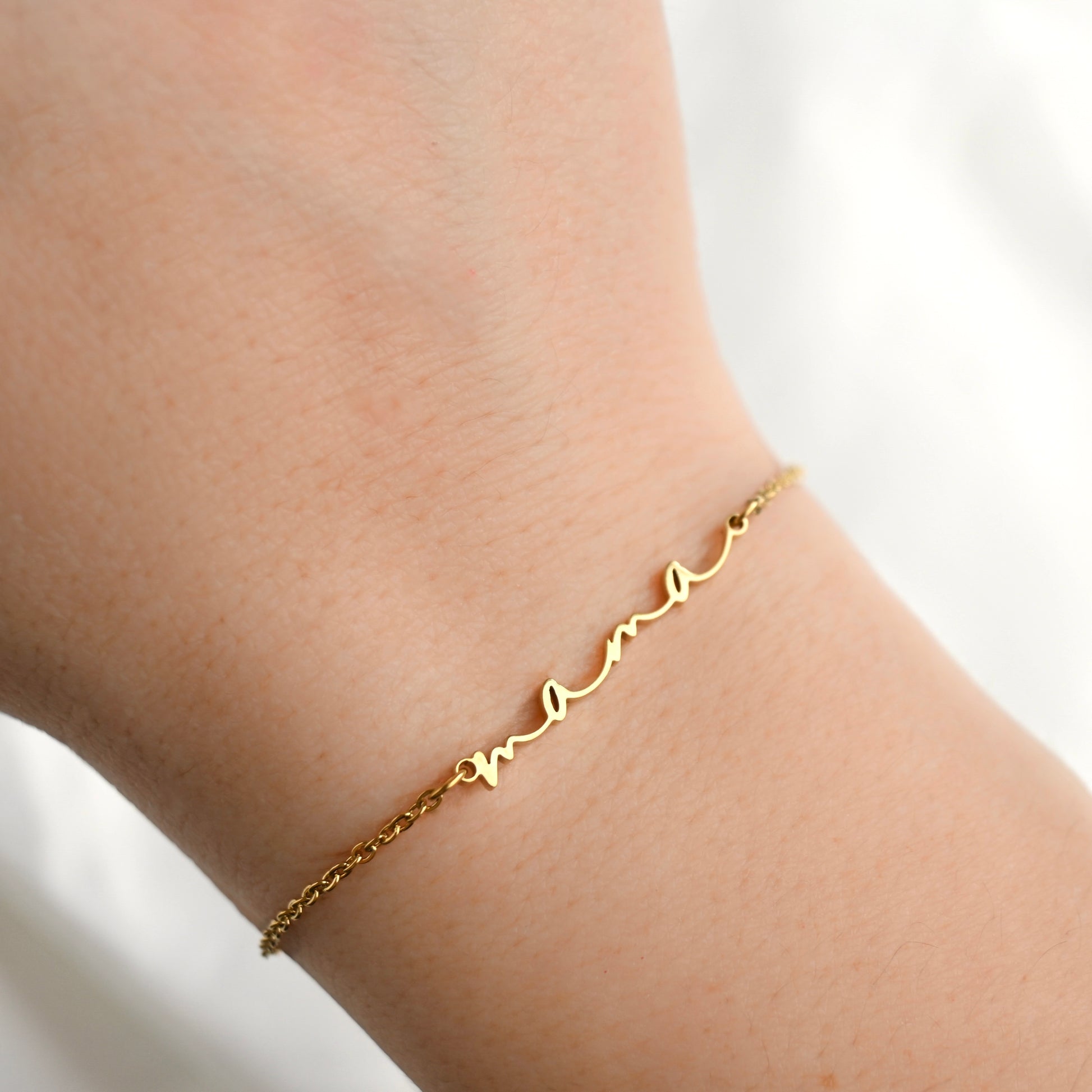 A delicate gold chain bracelet with a script name 'mama' engraved on it, shown worn on a person's wrist against a soft white background.