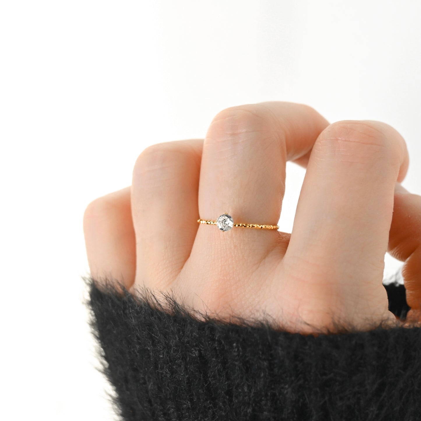 A delicate gold ring with a single round clear gemstone worn on a person's finger, shown against a bright white background with a black fuzzy sleeve visible at the bottom.
