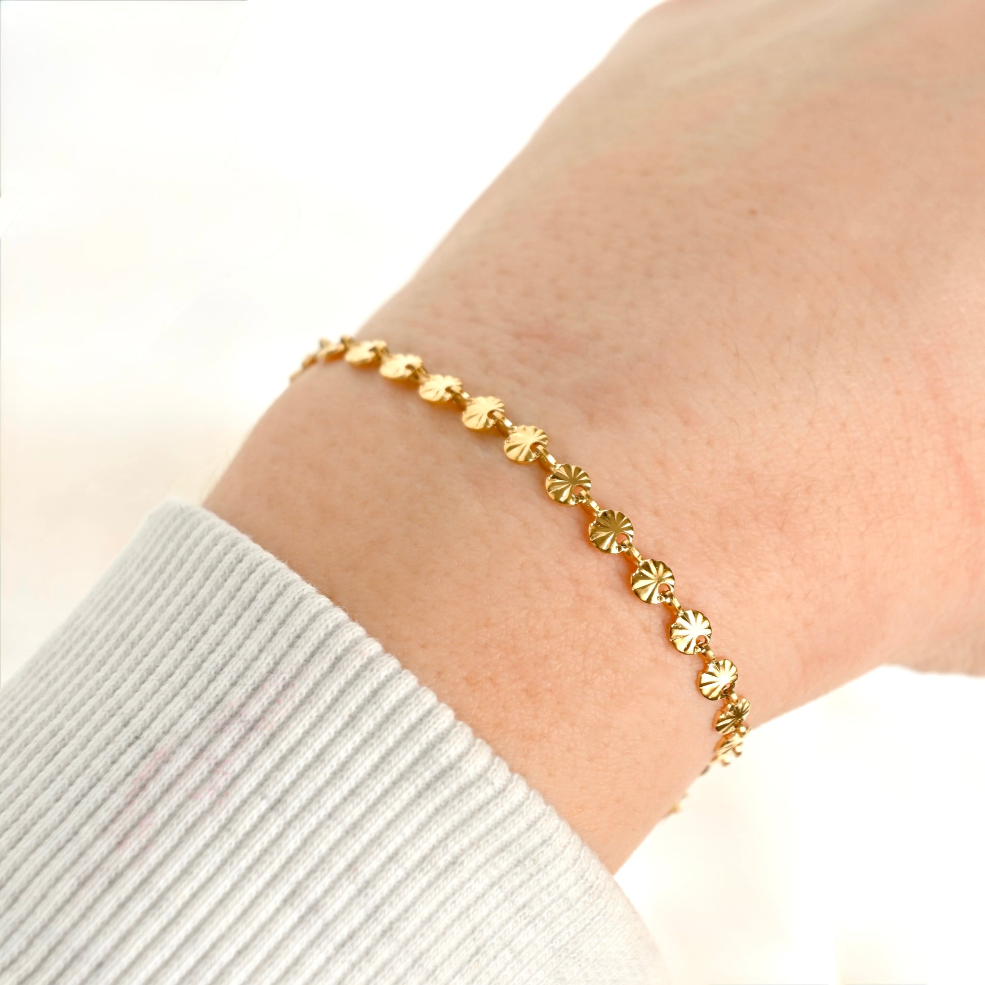 A delicate gold chain bracelet with small, textured sunburst links, shown worn on a person's wrist over a white ribbed long-sleeve cuff.