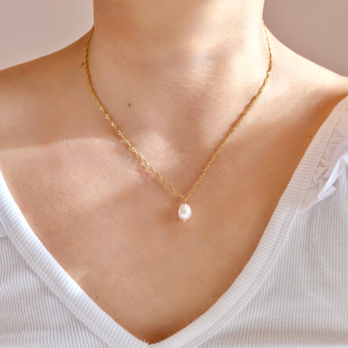 A delicate gold chain necklace with a single white freshwater pearl pendant, shown worn on a model's neck over a white ribbed top.