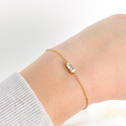 A delicate gold chain bracelet with a small rectangular clear gemstone centerpiece, shown worn on a person's wrist against a soft white background.