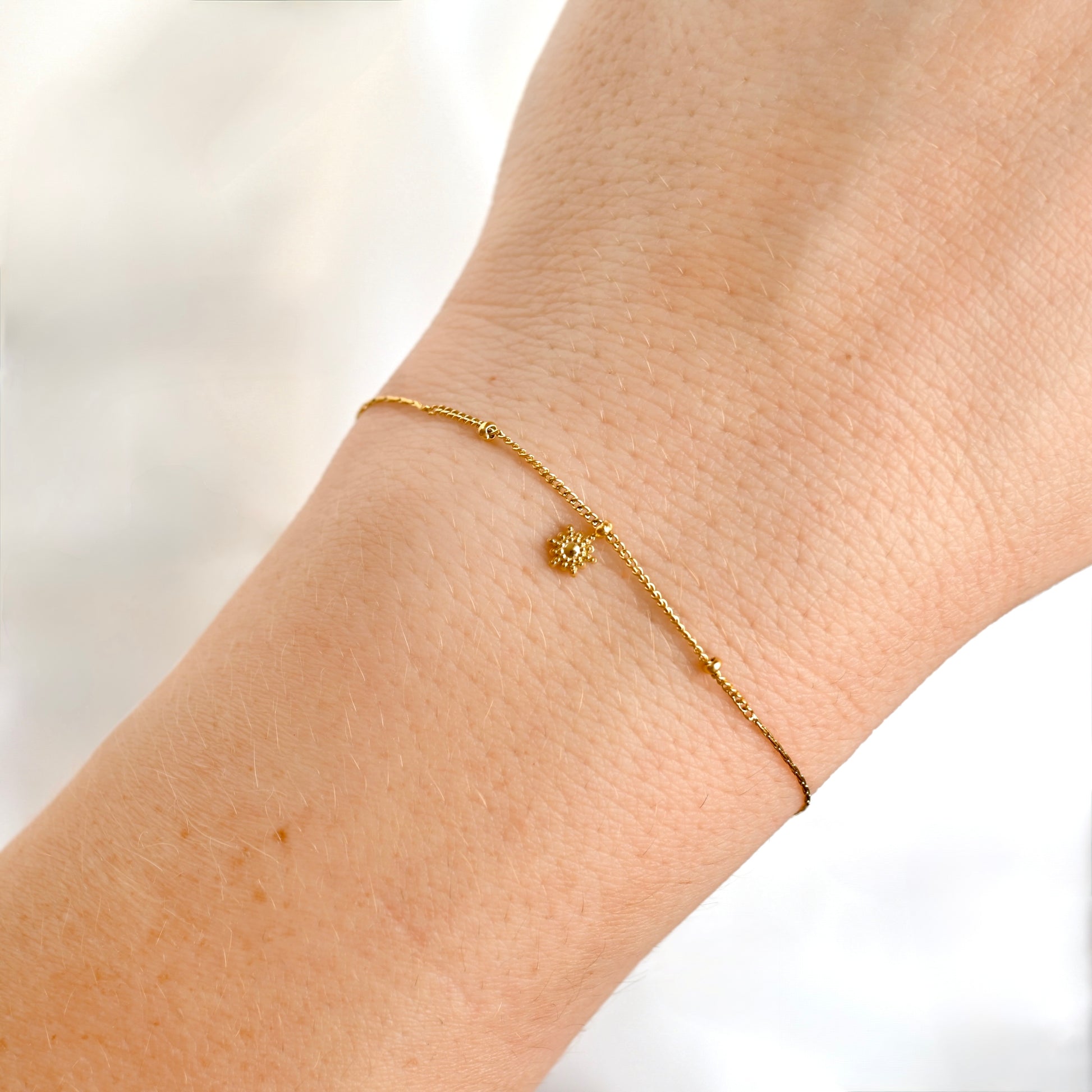 A delicate gold chain bracelet with a small sunburst charm, shown worn on a person's wrist against a soft, light background.