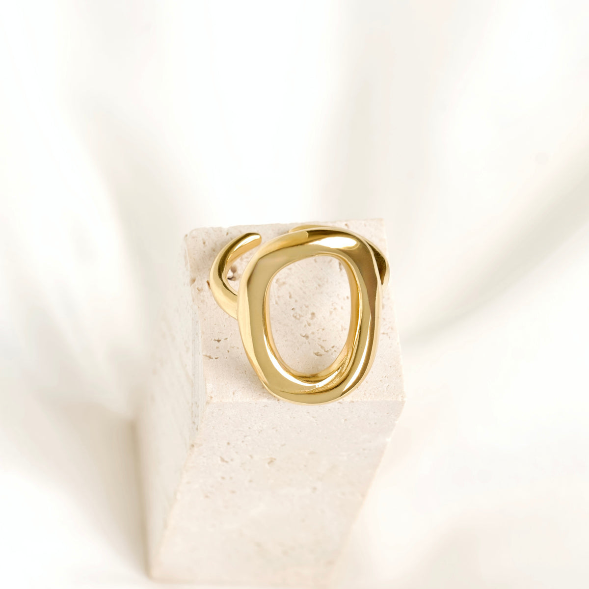 A polished gold ring with an abstract, interlocking loop design, displayed on a textured white stone block against a soft white fabric background.