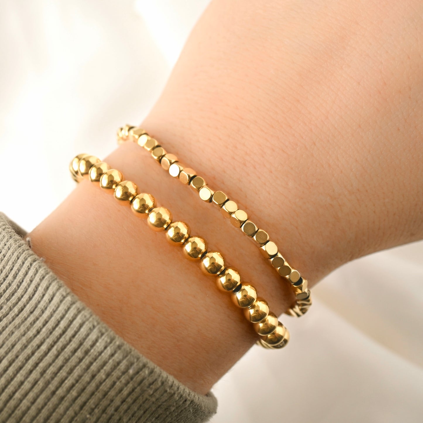 A stack of gold ball bead and cube bead bracelet on a wrist with a neutral background