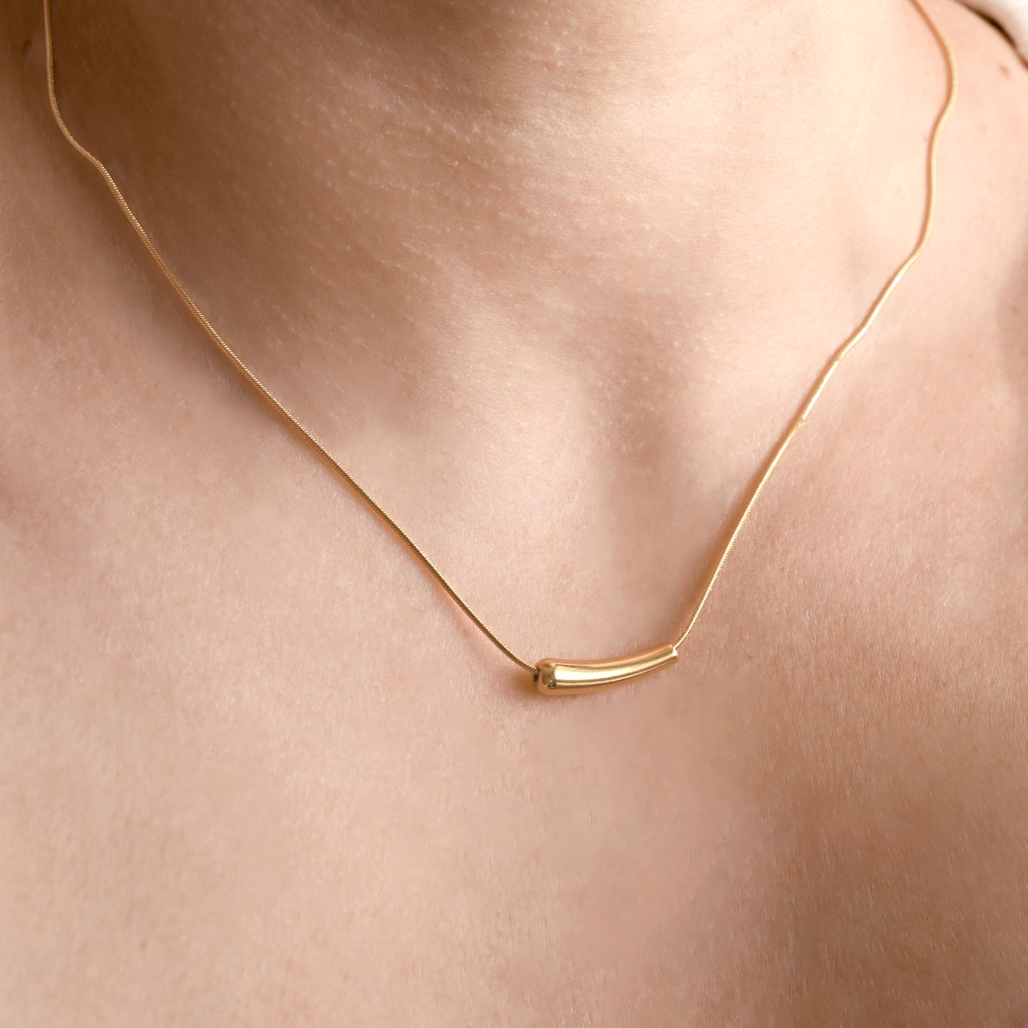 A close-up of a delicate gold chain necklace worn on a person's neck, featuring a small curved cylindrical pendant.