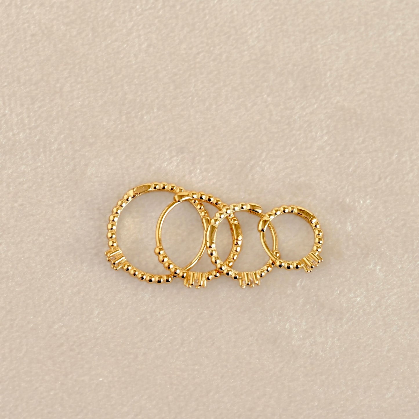 Top view of four gold-toned beaded hoop earrings arranged in a fan shape on a textured beige background.
