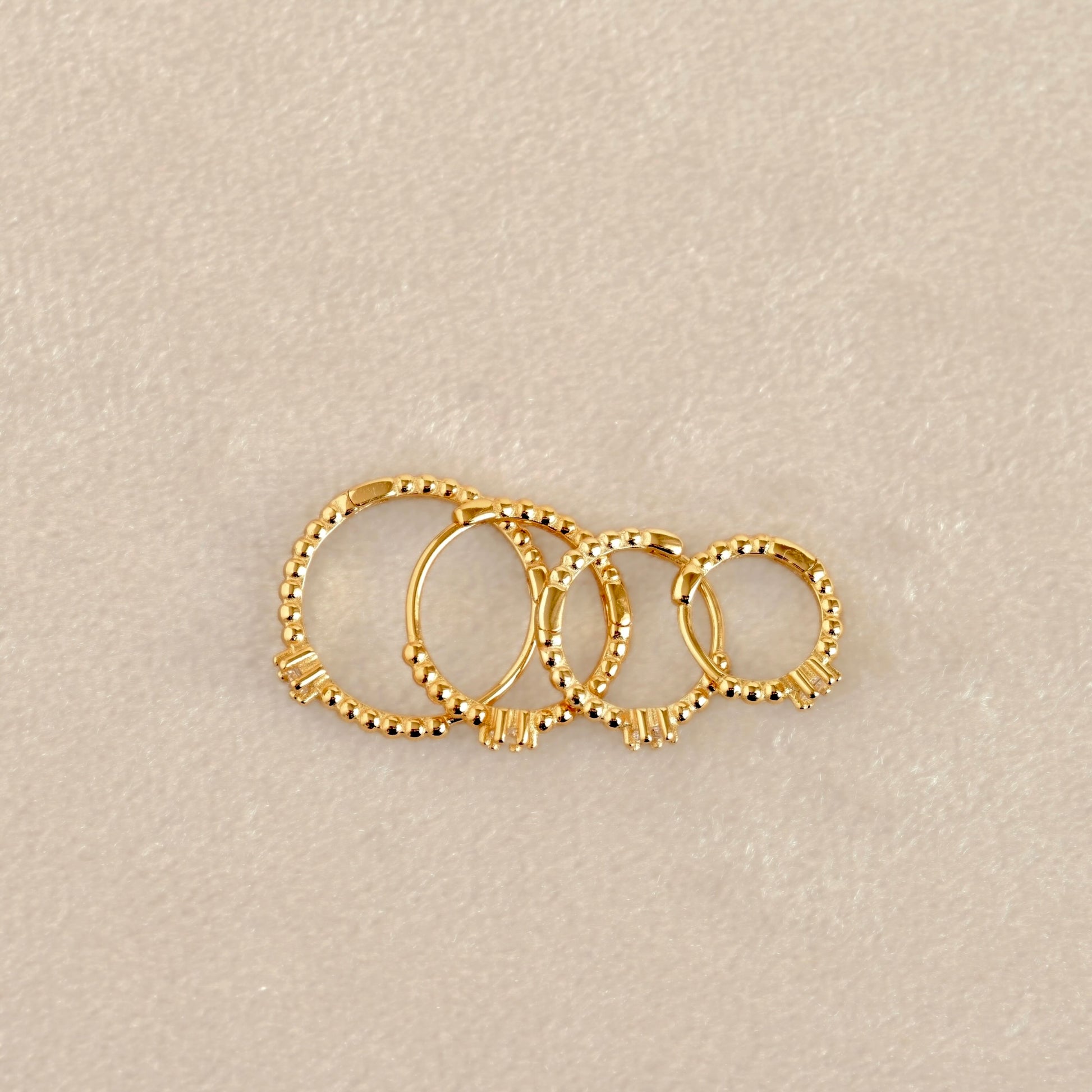 Top view of four gold-toned beaded hoop earrings arranged in a fan shape on a textured beige background.