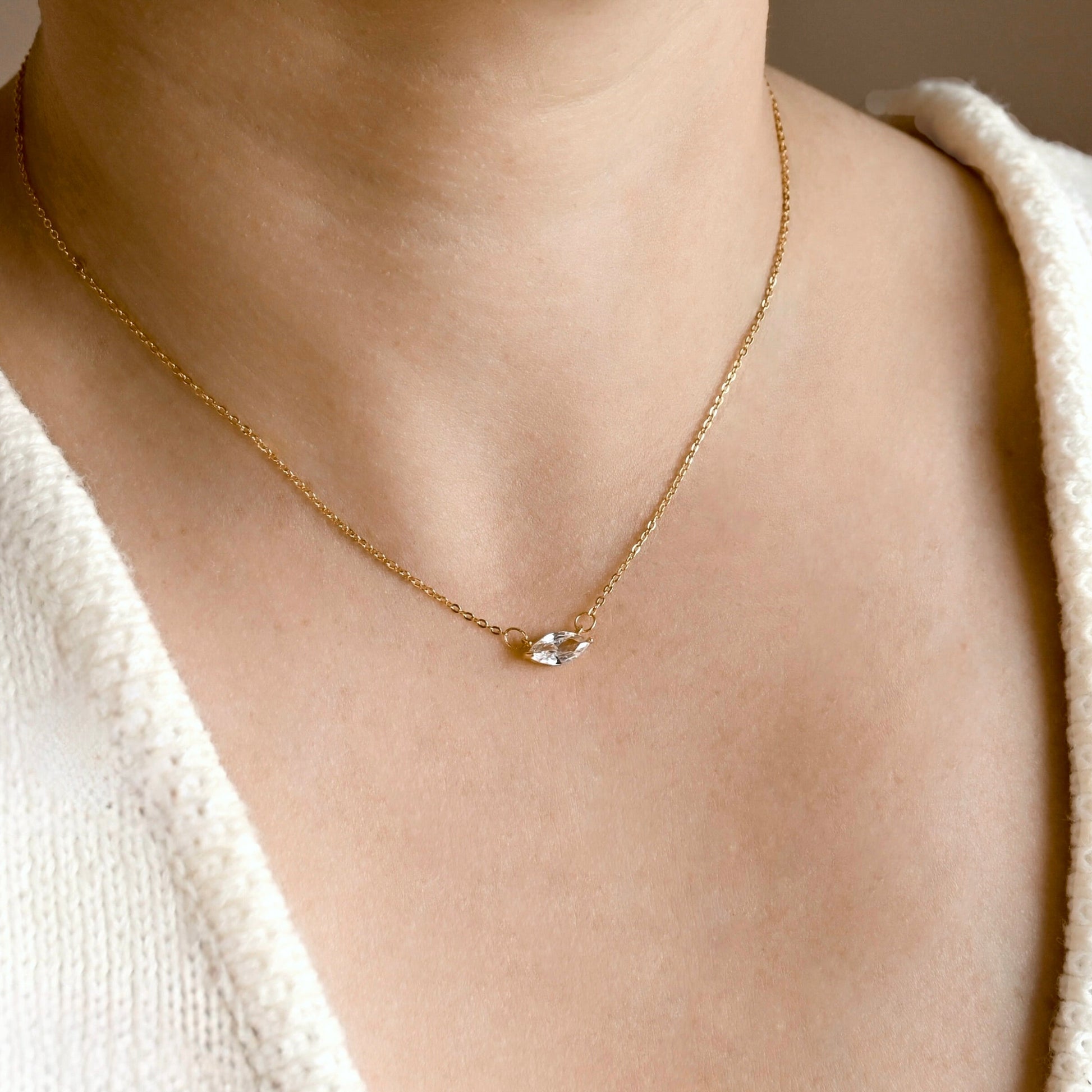A close-up of a person's neck and chest wearing a delicate gold chain necklace with a small clear marquise cut crystal pendant, paired with a white knit V-neck top.