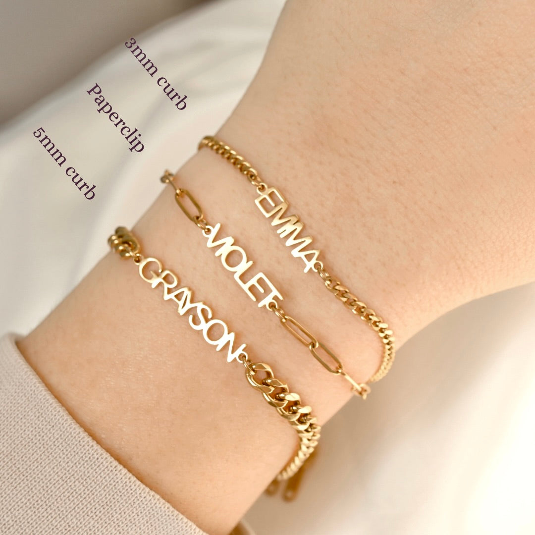 Three personalized gold name bracelets with different chain styles are shown stacked on a person's wrist. The bracelets feature the names 'EMMA', 'VIOLET', and 'GRAYSON' in gold-tone lettering.