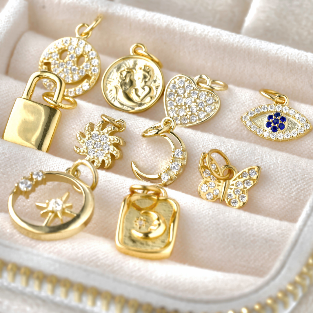 Collection of gold pendants  inlcuding smiley face, coin, heart, evil eye, padlock, sun, moon, butterfly and star on a textured beige surface