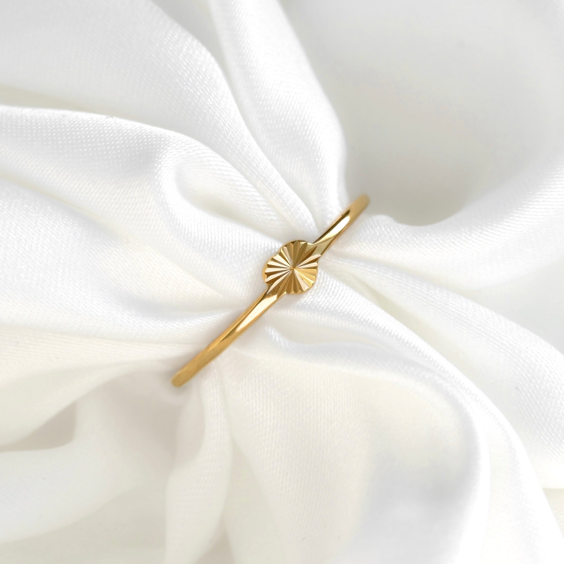 A delicate gold heart  ring with a sunburst or fan-shaped textured detail on the band, displayed on soft, draped white fabric.