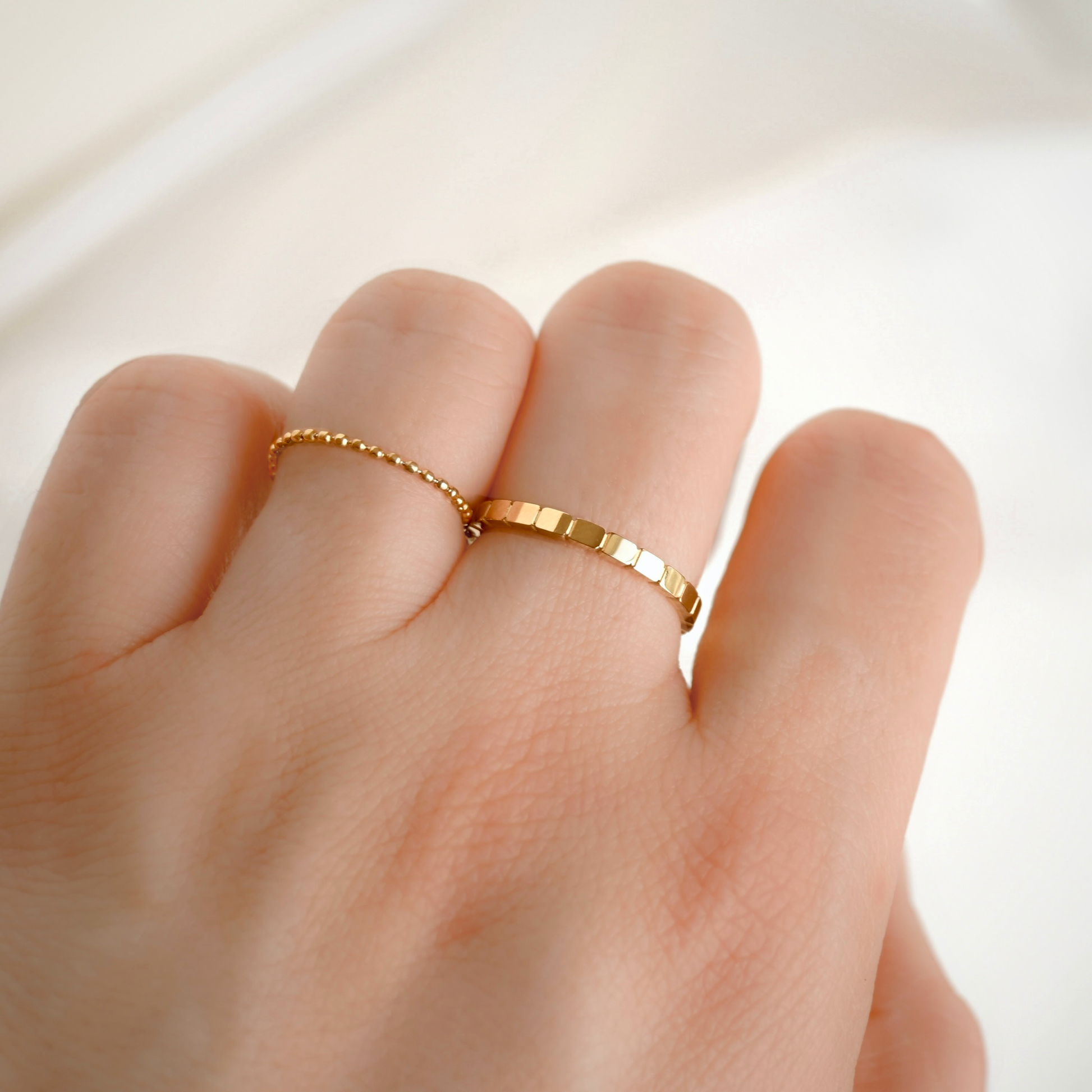 A close-up of a hand wearing two delicate gold rings, including one beaded chain ring and one flat textured band ring.