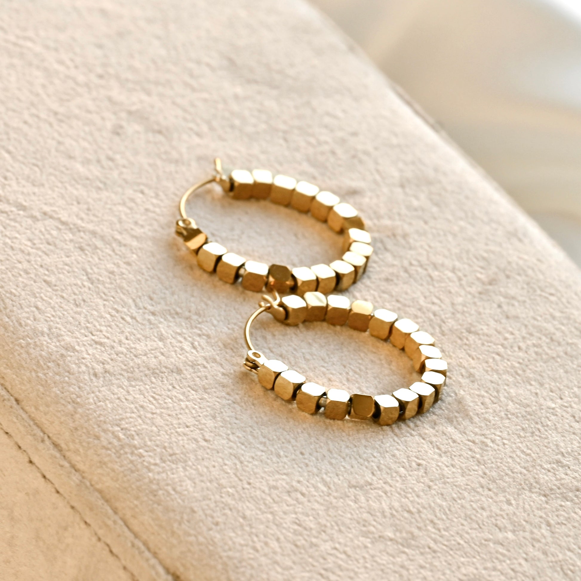 A pair of gold hoop earrings with a textured, faceted cube-like beaded design, resting on a beige fabric jewelry display.