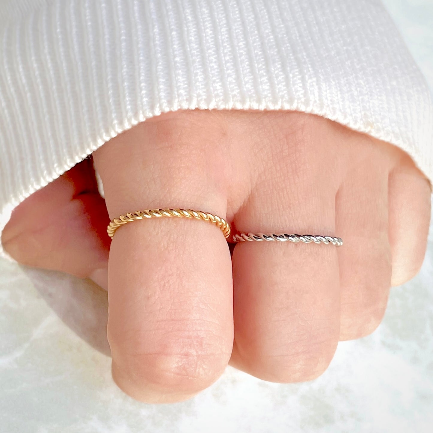 Close-up top view of a hand wearing two thin rope rings on a white fabric background