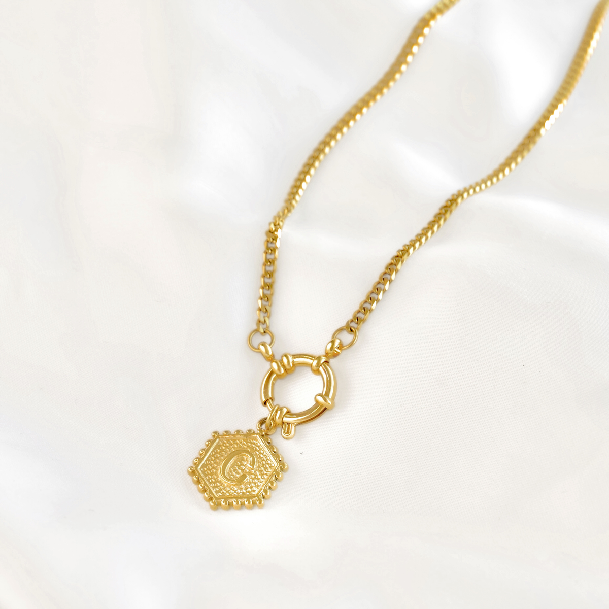 A gold chain necklace with a small hexagonal pendant featuring an engraved letter 'C', displayed on a soft white fabric background.