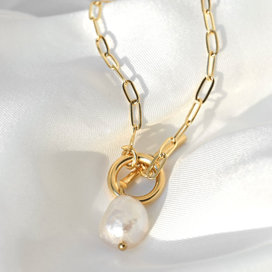 Gold paperclip chain necklace with a toggle clasp and a teardrop-shaped white baroque pearl pendant, displayed on a white satin fabric background.