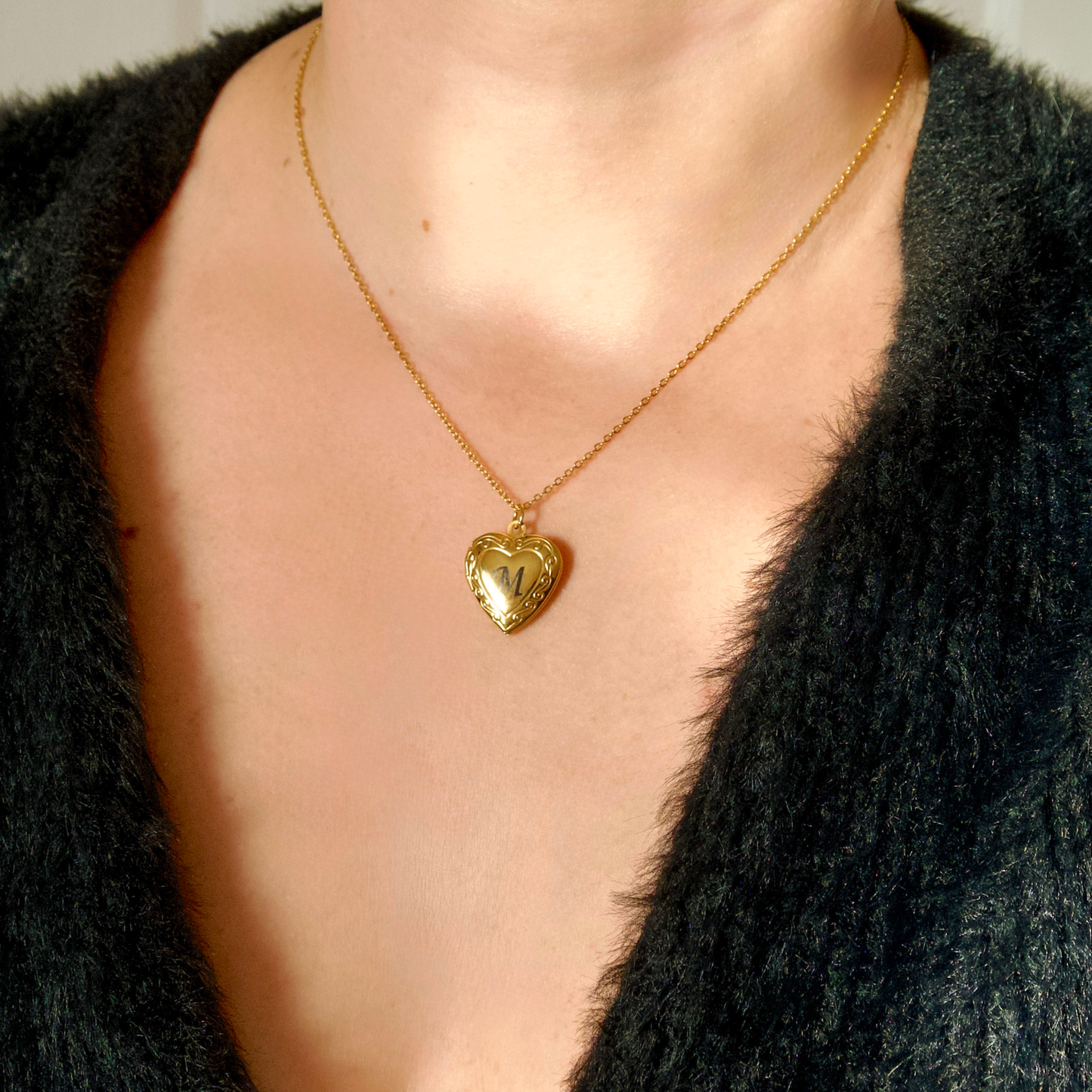 Gold heart-shaped necklace with 'M' initial on a person wearing a black garment.