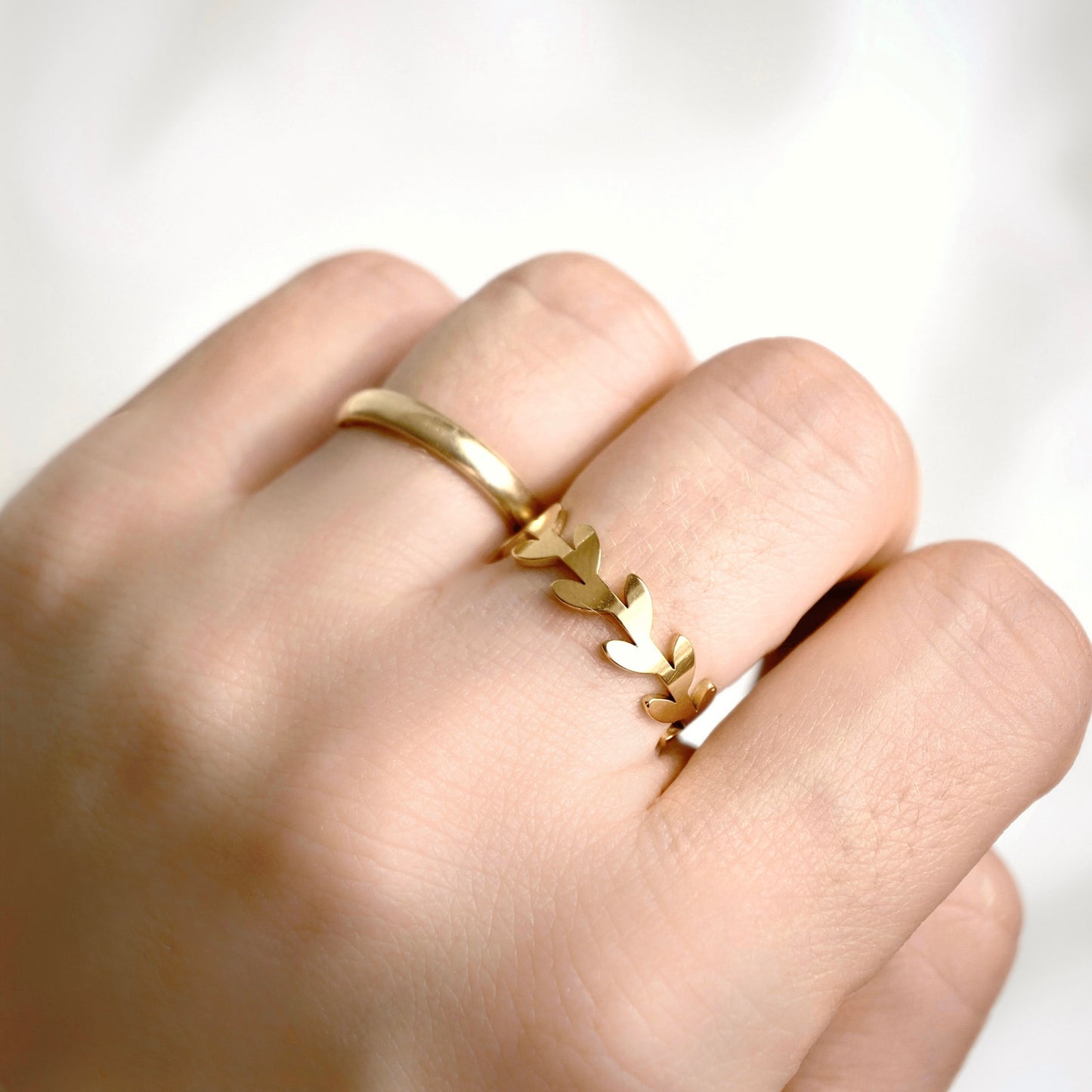 A close-up of a hand wearing a delicate gold leaf ring on one of the middle fingers, set against a soft, out-of-focus white background.