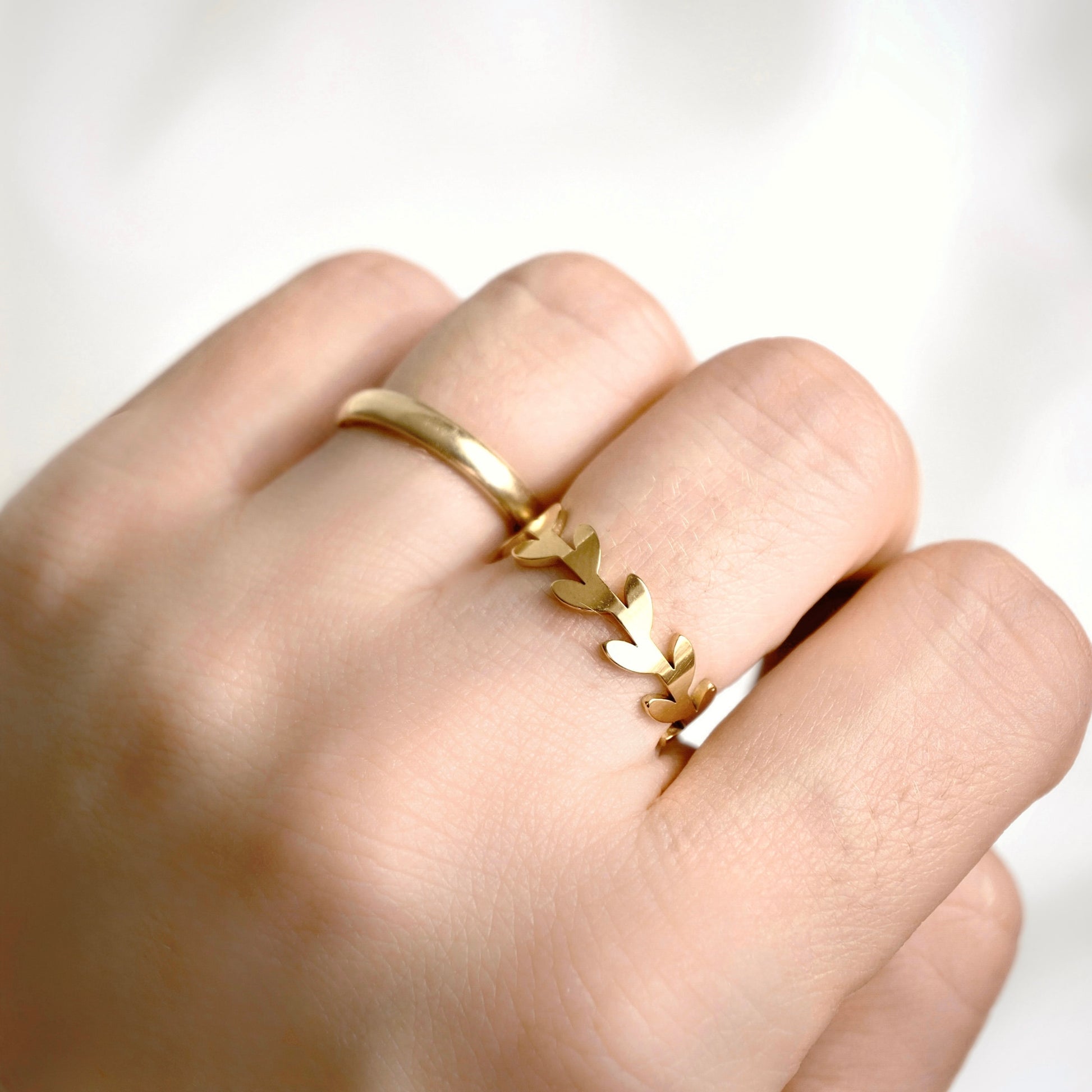 A close-up of a hand wearing a delicate gold leaf ring on one of the middle fingers, set against a soft, out-of-focus white background.