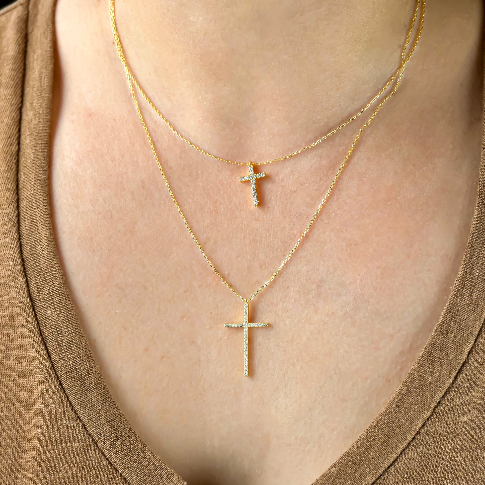 Two gold cross necklaces, one with medium size and one with large cross charm on a person wearing a brown top.