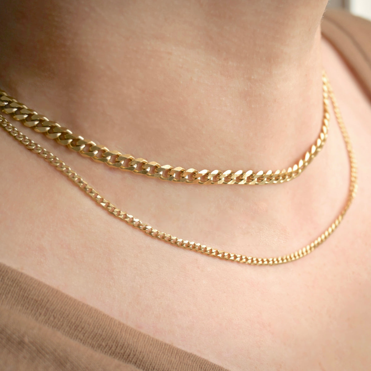 Two gold-toned chain necklaces, one chunky curb link and one delicate curb chain, worn layered on a person's neck.