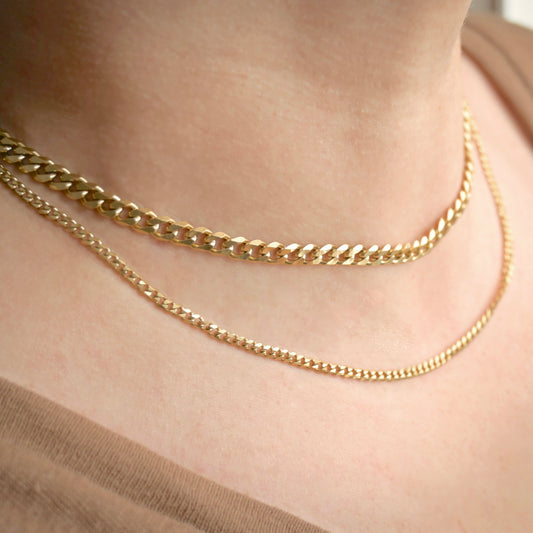 Two gold-toned chain necklaces, one chunky curb link and one delicate curb chain, worn layered on a person's neck.